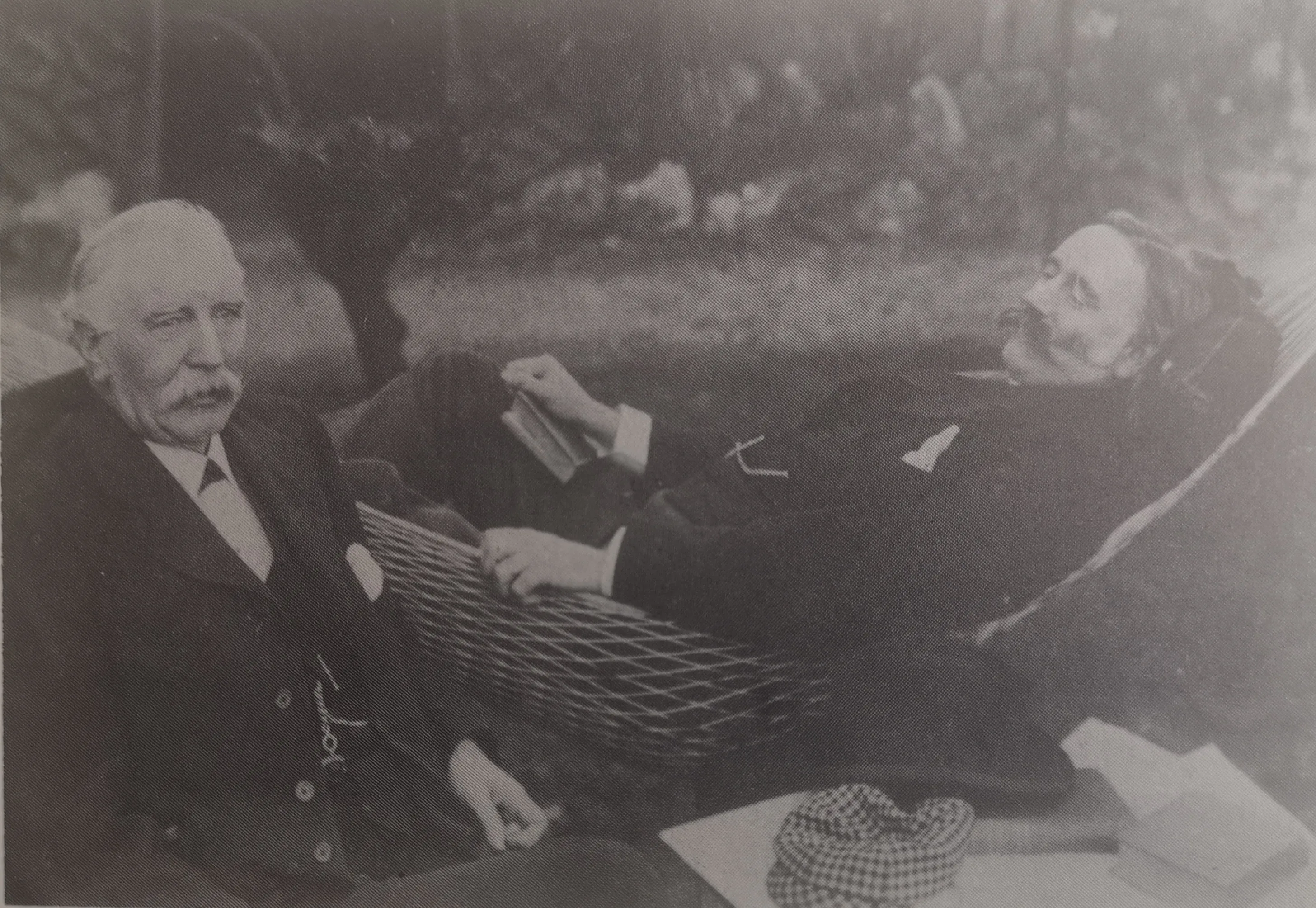 A man sits at a folding table beside another man reclining in a hammock outside